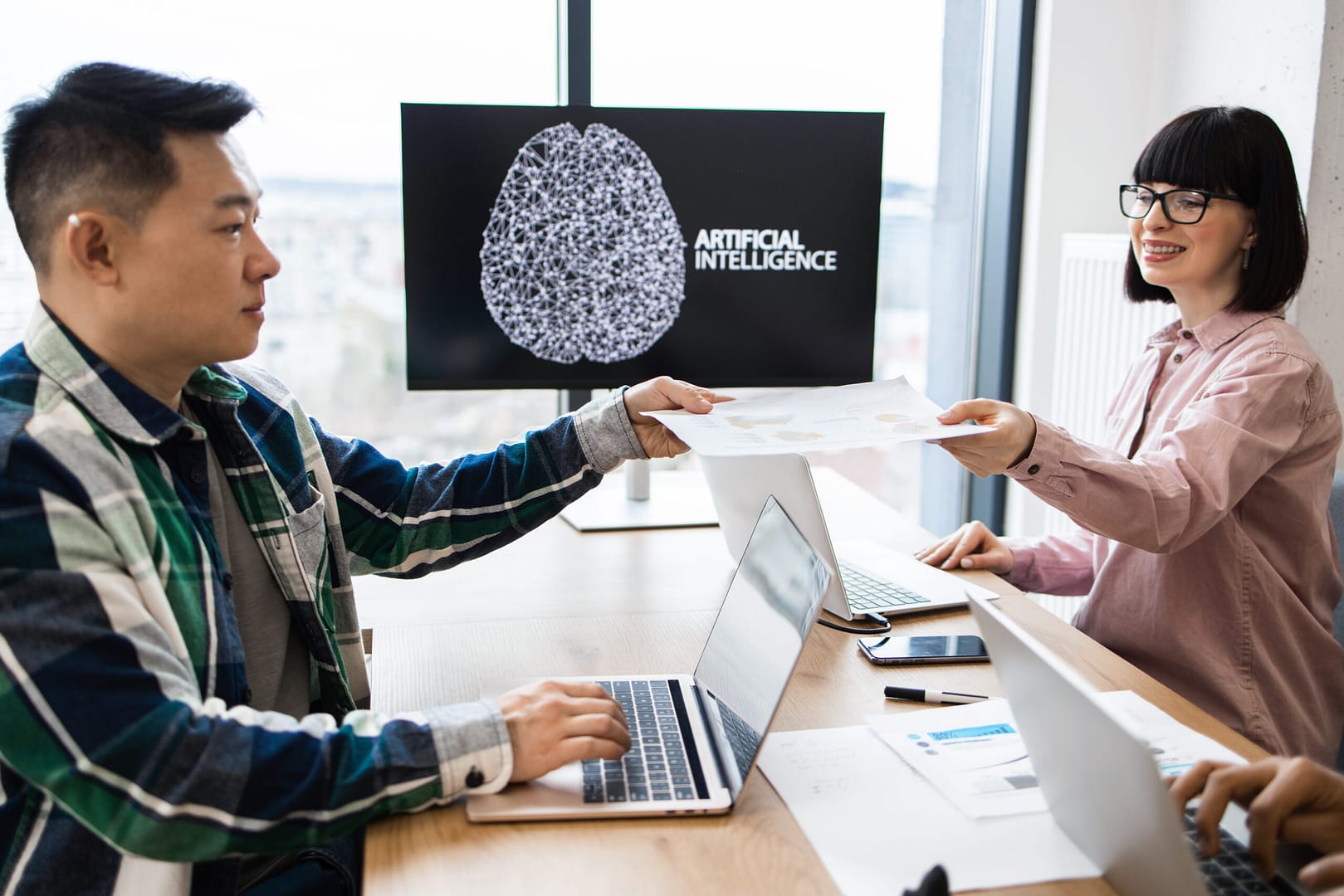 Membros da equipe colaborando em um projeto de inteligência artificial em um ambiente de escritório moderno. Duas pessoas compartilhando documentos e ideias enquanto trabalham em laptops, com foco em tecnologia inovadora.