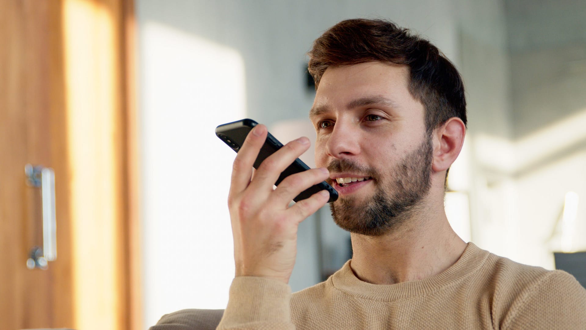 Homem sorridente gravando uma mensagem de áudio no telefone. Homem atraente com telefone no sofá