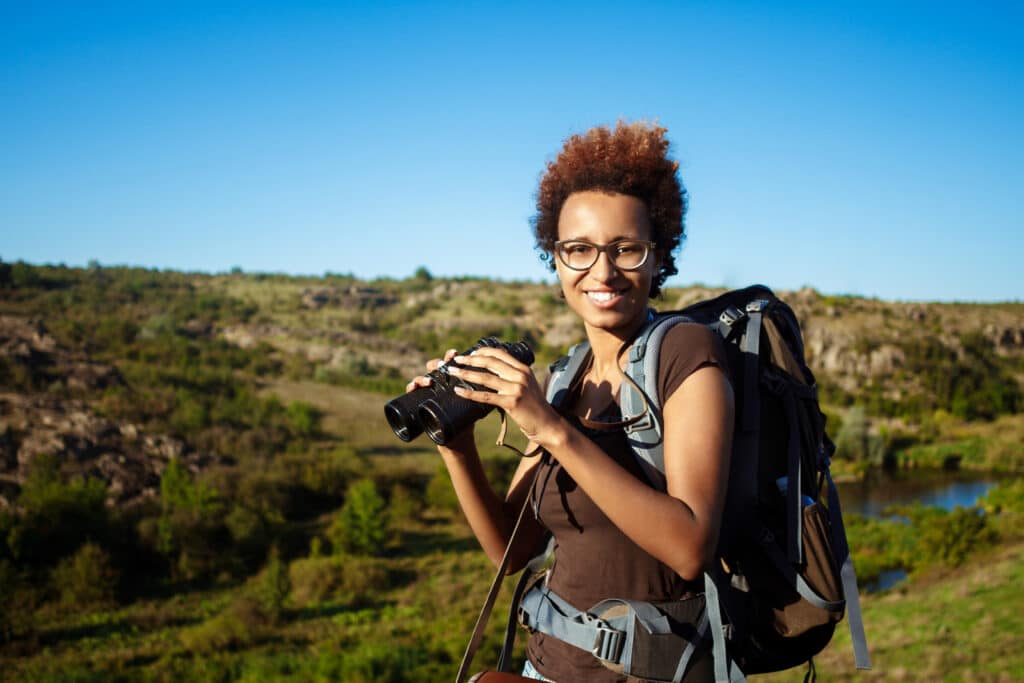 Young beautiful african girl with backpack smiling, looking at camera, holding binoculars, canyon background.