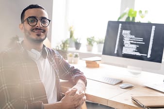 Retrato de jovem programador positivo do Oriente Médio em óculos sentado com as mãos entrelaçadas na mesa com um computador poderoso